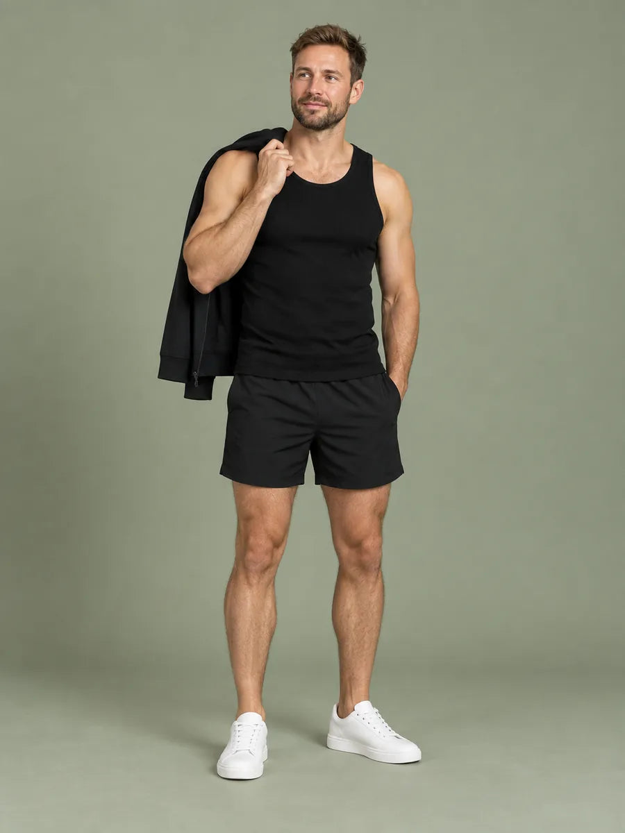 Mann in schwarzem Ripp-Tanktop und schwarzen Shorts, ein schwarzes Half-Zip über der Schulter, vor graugrünem Studiohintergrund