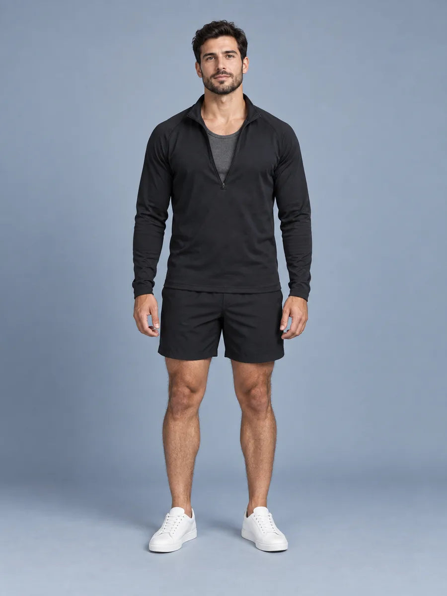 Mann in offenem schwarzem Half-Zip über einem anthrazitfarbenen Ripp-Tanktop und schwarzen Shorts, vor blaugrauem Studiohintergrund
