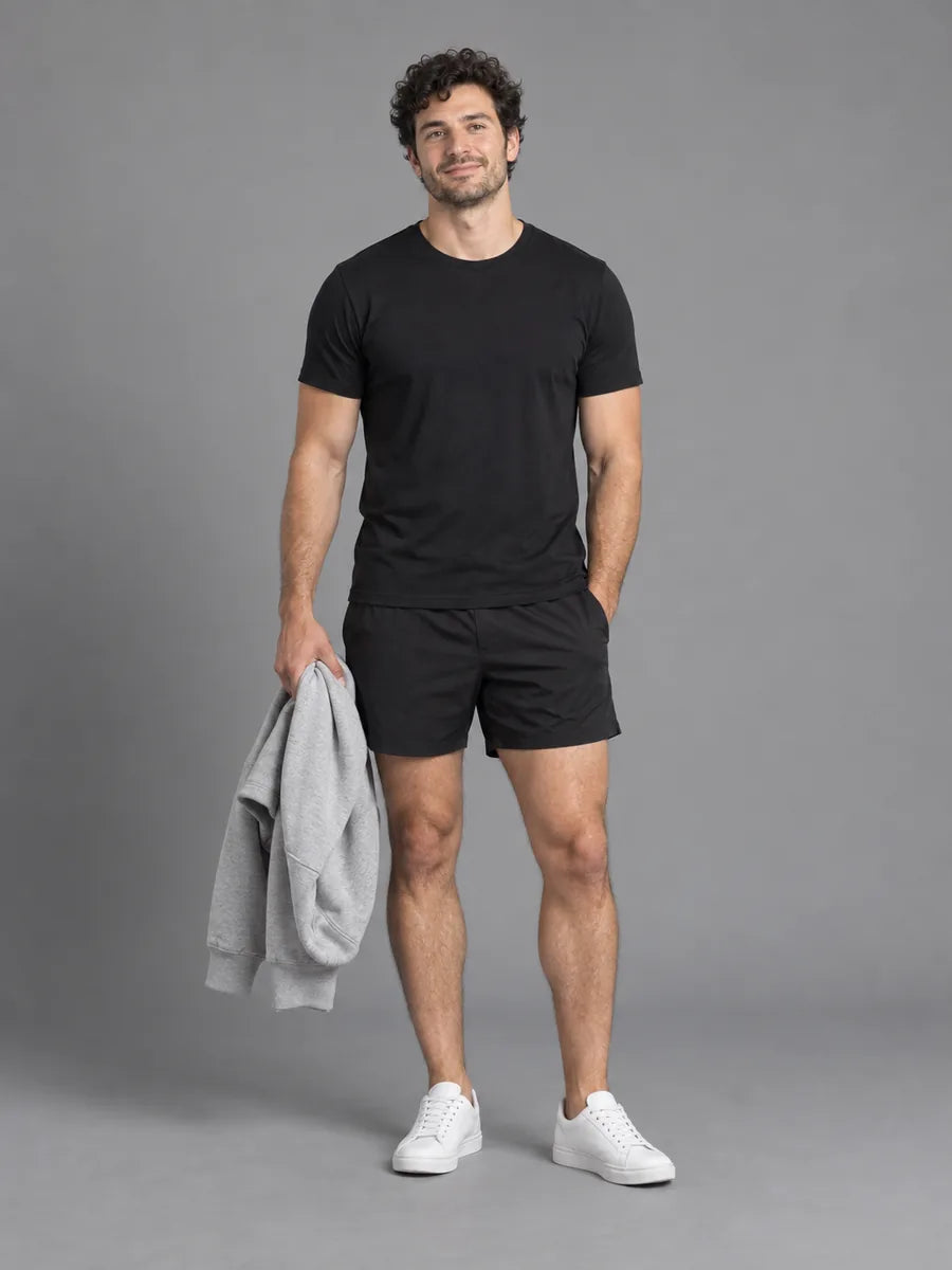 Mann in schwarzem Slim-Fit-T-Shirt und schwarzen Shorts, einen hellgrauen Hoodie in der Hand, vor grauem Studiohintergrund