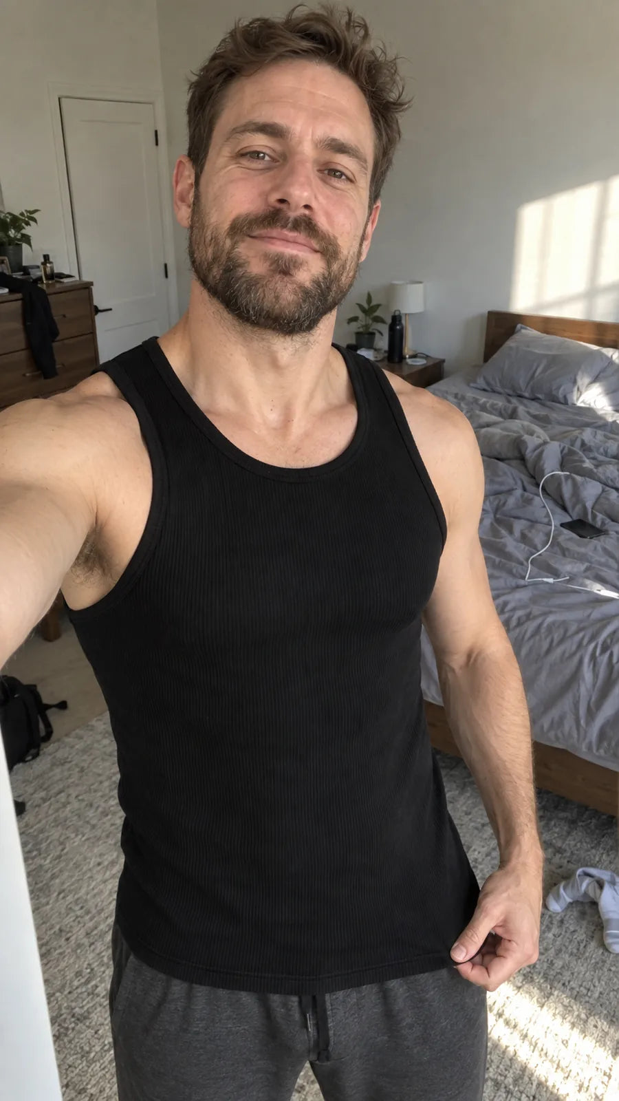 Ein Mann in schwarzem Ripp-Tanktop zieht im Schlafzimmer den Saum nach unten, um die Länge an der Hüfte zu zeigen.
